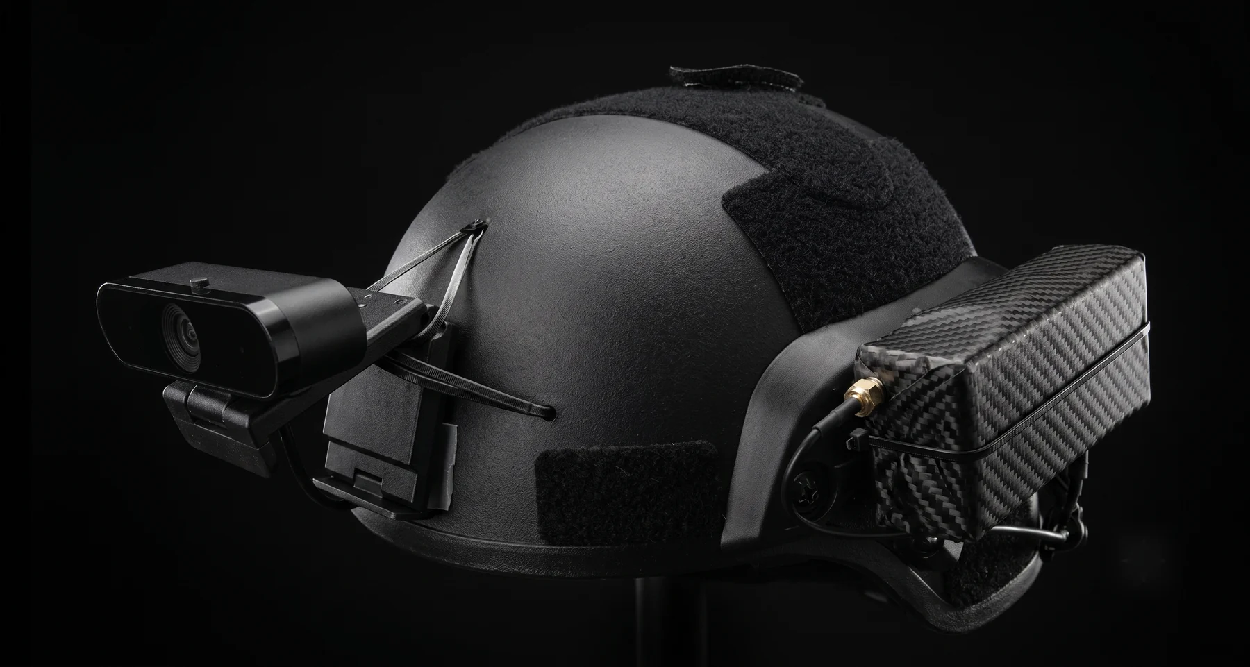 A ballistic helmet carrying a camera on a side bracket and a carbon-wrapped
                control box on the opposite rail, with an antenna connector